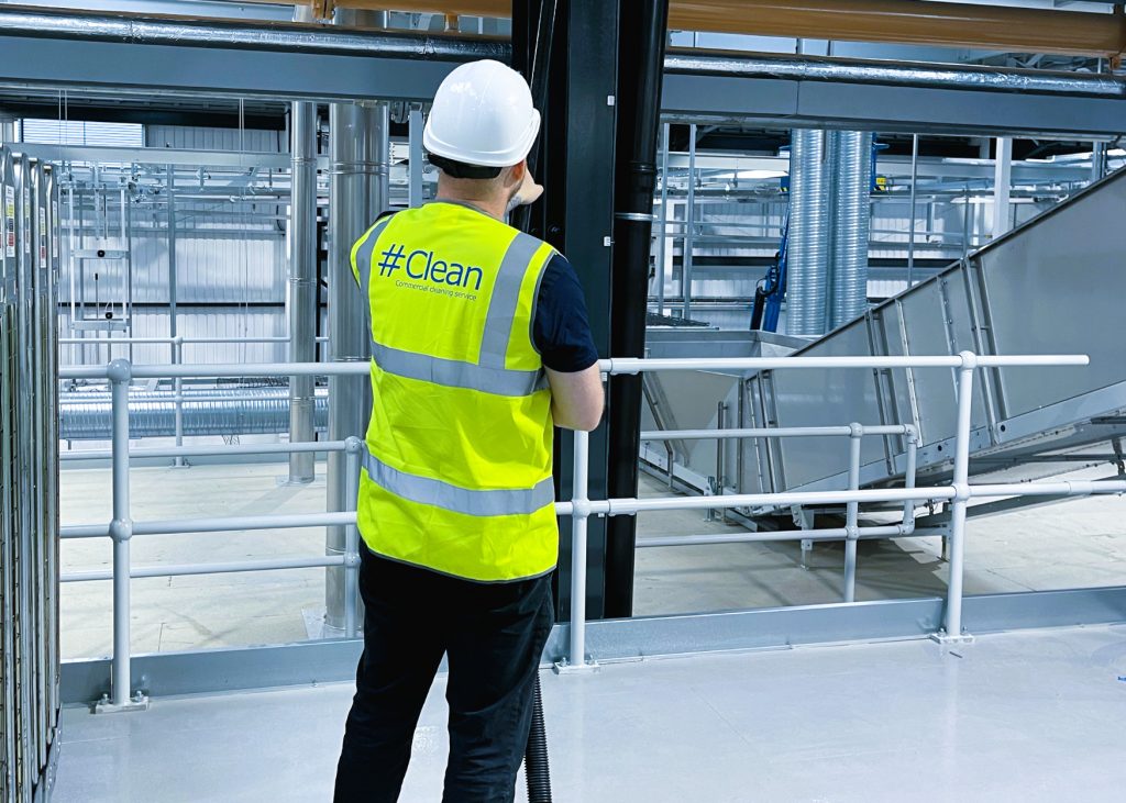 Hashtag Clean worker in a high-visibility vest and hard hat performing cleaning duties in a large industrial facility with exposed ducting and metal structures.