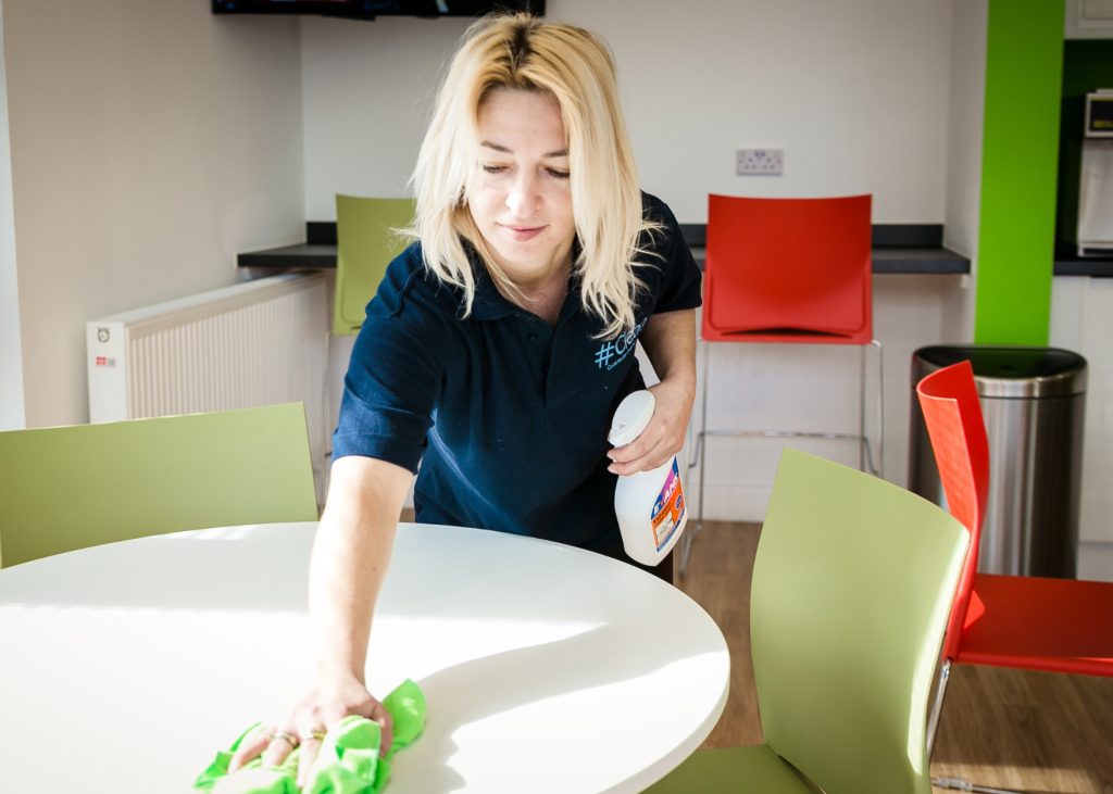 Hashtag Clean staff member wiping down a round table in a modern office kitchen or break room using a green cloth and disinfectant spray.