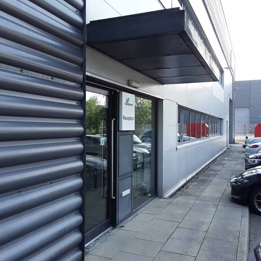 The exterior of a commercial building with a glass reception door, an awning, and parked cars at the side.