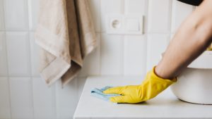 A cleaner wearing a bright yellow glove wipes a white countertop with a blue cleaning cloth near a white sink and tiled wall.