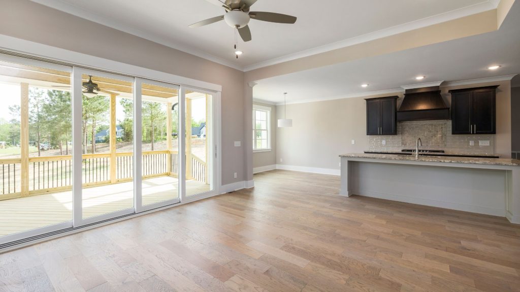 A bright, empty open-plan rental property interior featuring polished hardwood floors, a fitted kitchen with dark cabinets, large sliding glass doors leading to a decked balcony, and a ceiling fan — ready for end of tenancy inspection.
