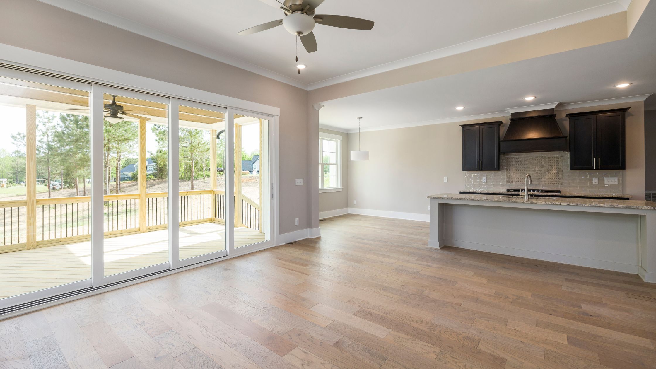 A bright, empty open-plan rental property interior featuring polished hardwood floors, a fitted kitchen with dark cabinets, large sliding glass doors leading to a decked balcony, and a ceiling fan — ready for end of tenancy inspection.