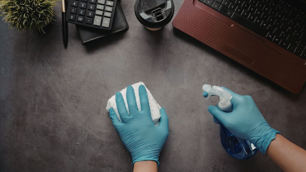 An overhead view of a professional cleaner wearing blue latex gloves wiping down a grey office desk surface with a white cloth and blue spray bottle, with a laptop, calculator, and coffee cup visible in the background.
