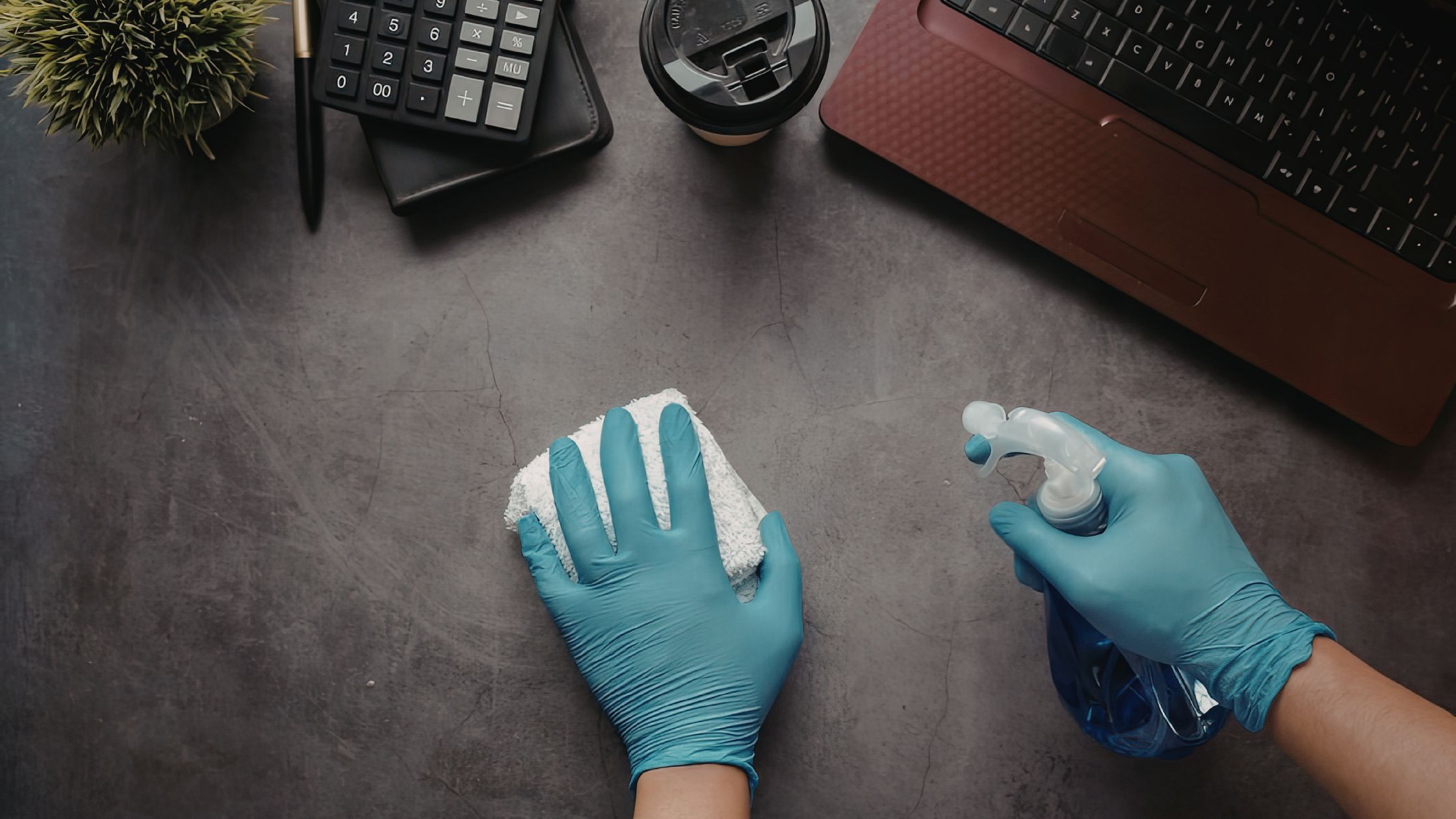 An overhead view of a professional cleaner wearing blue latex gloves wiping down a grey office desk surface with a white cloth and blue spray bottle, with a laptop, calculator, and coffee cup visible in the background.