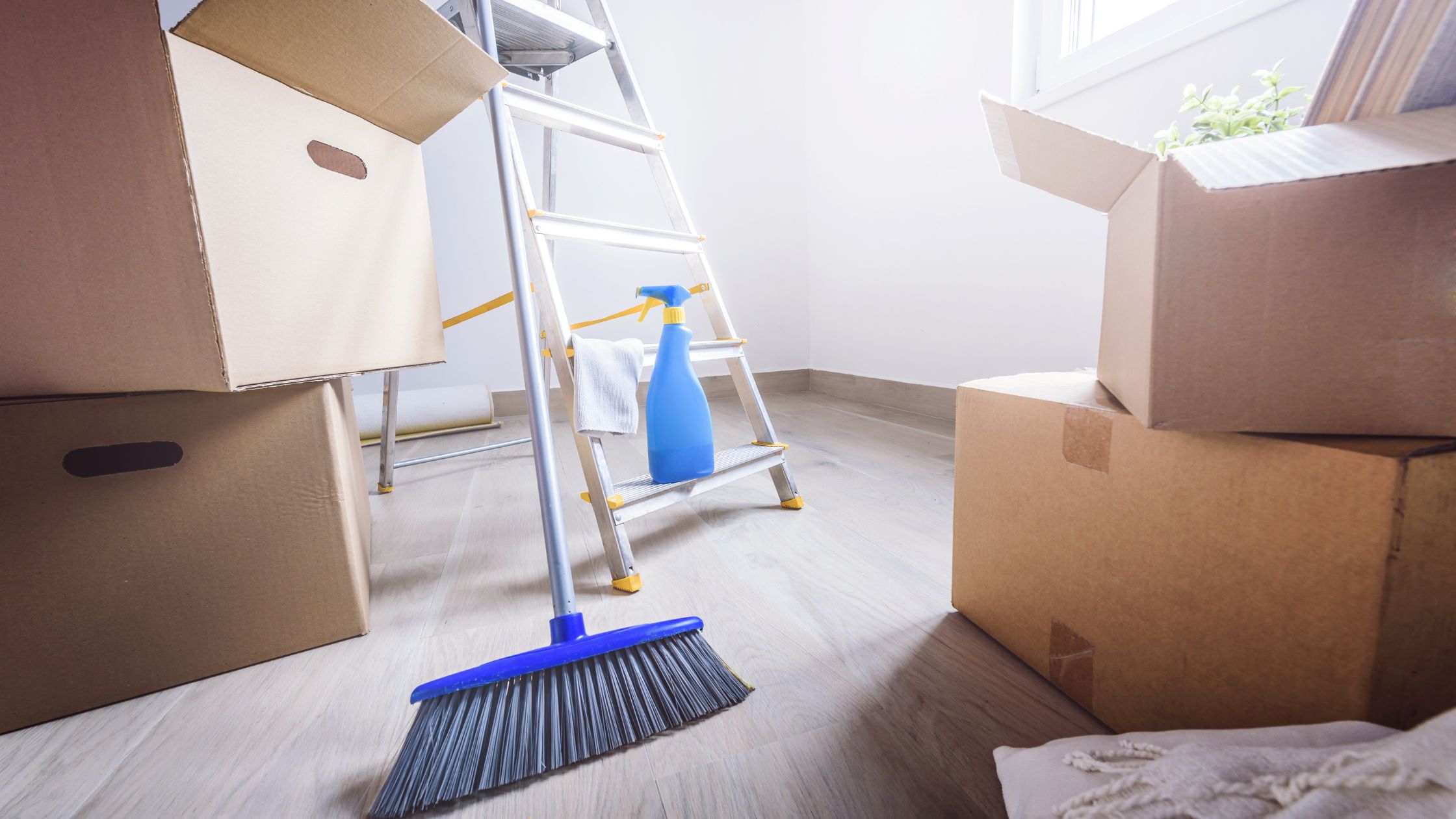 A broom, blue spray bottle, and white cloth resting against a step ladder surrounded by open cardboard moving boxes on a light wood floor in an empty rental property being prepared for an end of tenancy clean.
