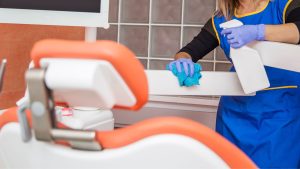 A professional cleaner in a blue uniform and purple gloves spraying and wiping a surface next to a dental treatment chair in a clinical setting.