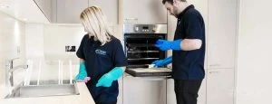 Two Hashtag Clean staff members in blue uniforms clean a modern kitchen, one scrubbing the sink, the other cleaning an oven.